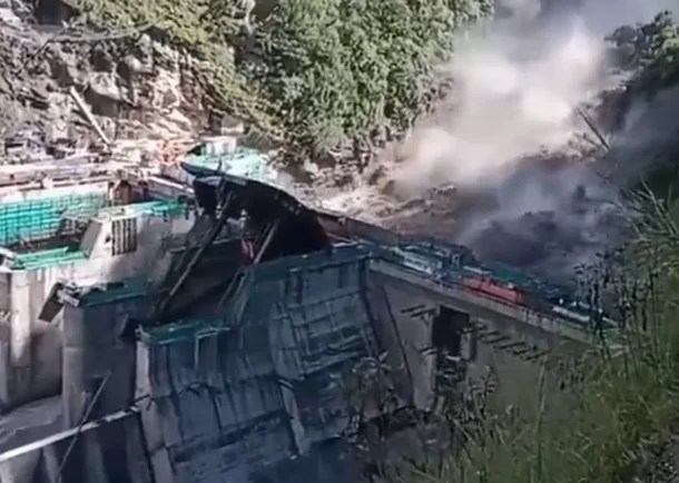 A damaged dam structure with debris and dust cloud from a landslide in the background, surrounded by trees.